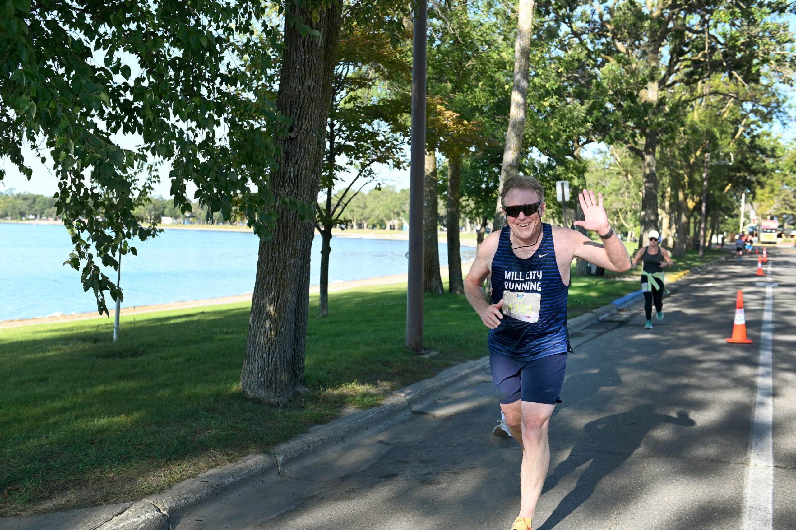 A male racer, wearing sunglasses, waves to camera as he runs.